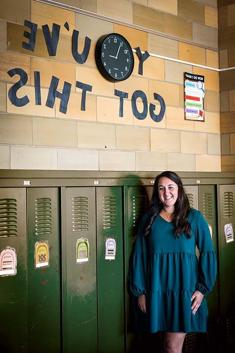 梅根·凯利, 穿着蓝色的裙子, 站在一所小学走廊上的一排深绿色储物柜前.