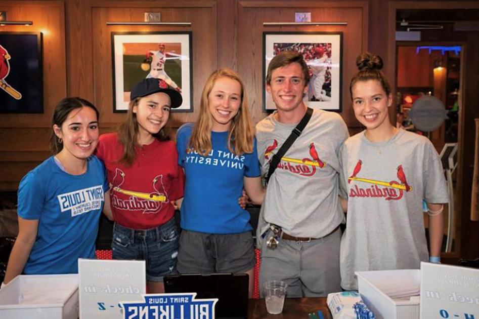 A group of five students wearing 博彩网址大全 and St. Louis Cardinals T-shirts pose in a wood-paneled room with framed photos of Cardinals baseball players on the wall.
