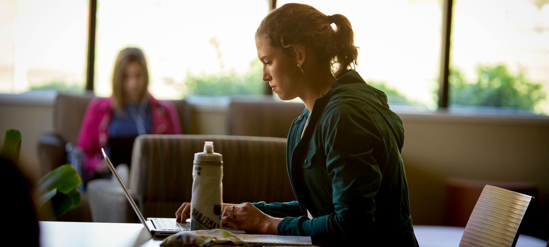 一个学生在桌子旁学习，桌子上放着水瓶和笔记本电脑，另一个学生坐在窗户前的背景中.